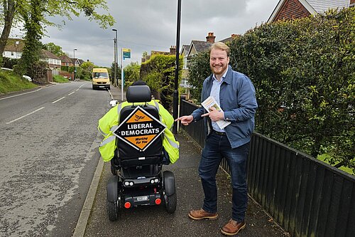 A person in a high-visibility jacket on a motorized wheelchair with a Liberal Democrats sign in a residential street, and a smiling man standing next to them holding leaflets.